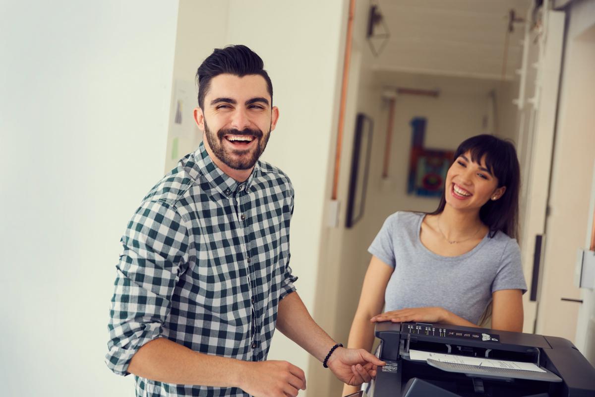 A smiling man using a multifunction copier while a coworker looks on.